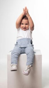 pexels photo 15443078 15443078 Cute child wearing blue overalls and white shoes, clapping with joy in a studio shoot.